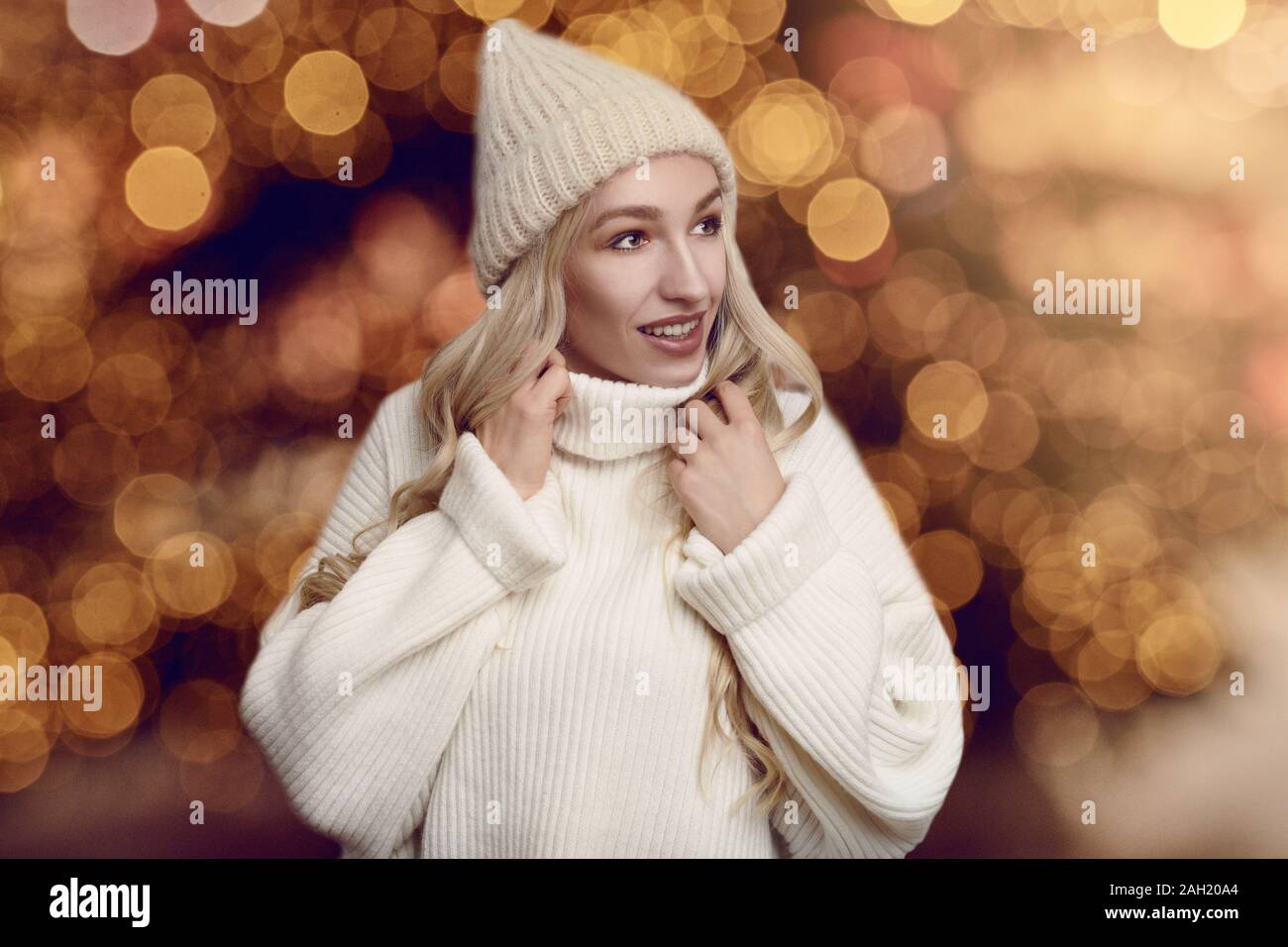 Schöne junge blonde Frau in Weiß gestrickte Pullover und Winter Hut, von der Kamera schauen und lächeln, stehend gegen warme Gelb leuchten mit b verschwommen Stockfoto