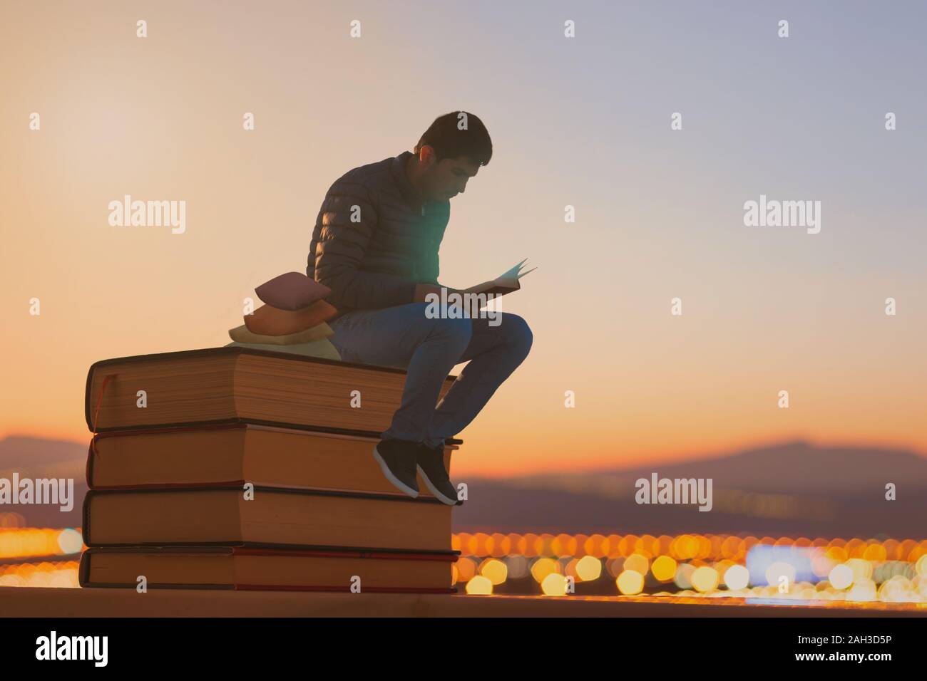 Ein Junge mit einem Buch auf einem Stapel Bücher im Freien mit dem Blick auf die Berge bei Sonnenuntergang. Stockfoto