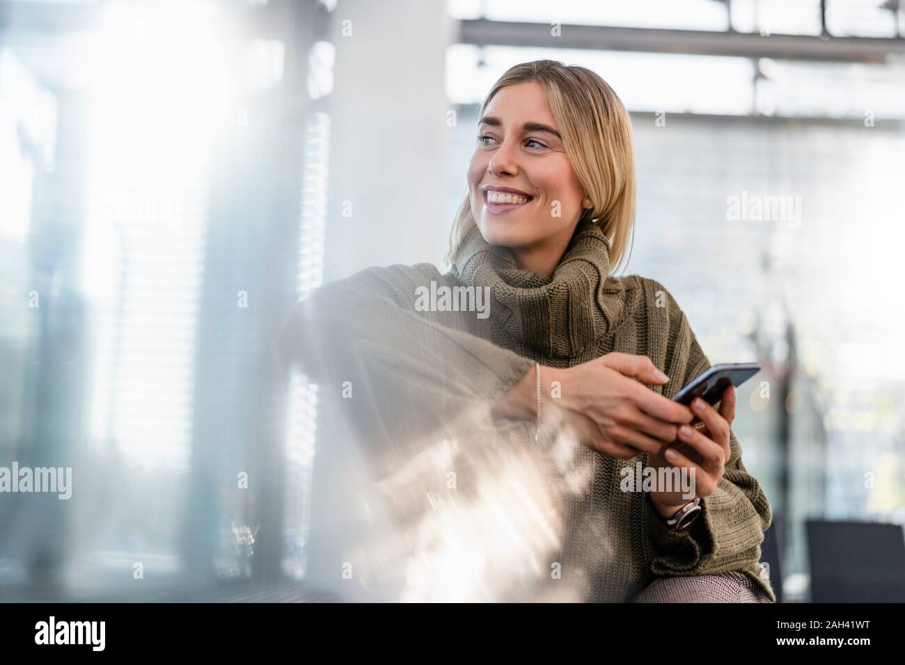 Lächelnde junge Frau mit Handy sitzen im Wartebereich um suchen Stockfoto