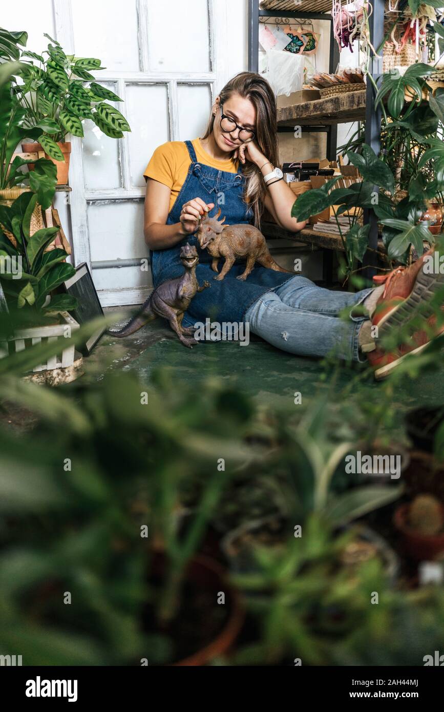 Junge Frau mit Dinosaurier Figur sitzen auf dem Boden in einem kleinen Laden mit Pflanzen Stockfoto