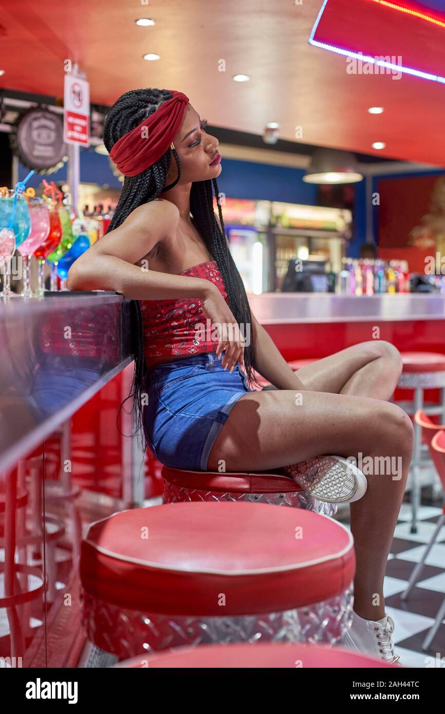 Junge Frau mit geflochtenem Frisur sitzt auf einem Bar auf dem roten Stuhl Stockfoto