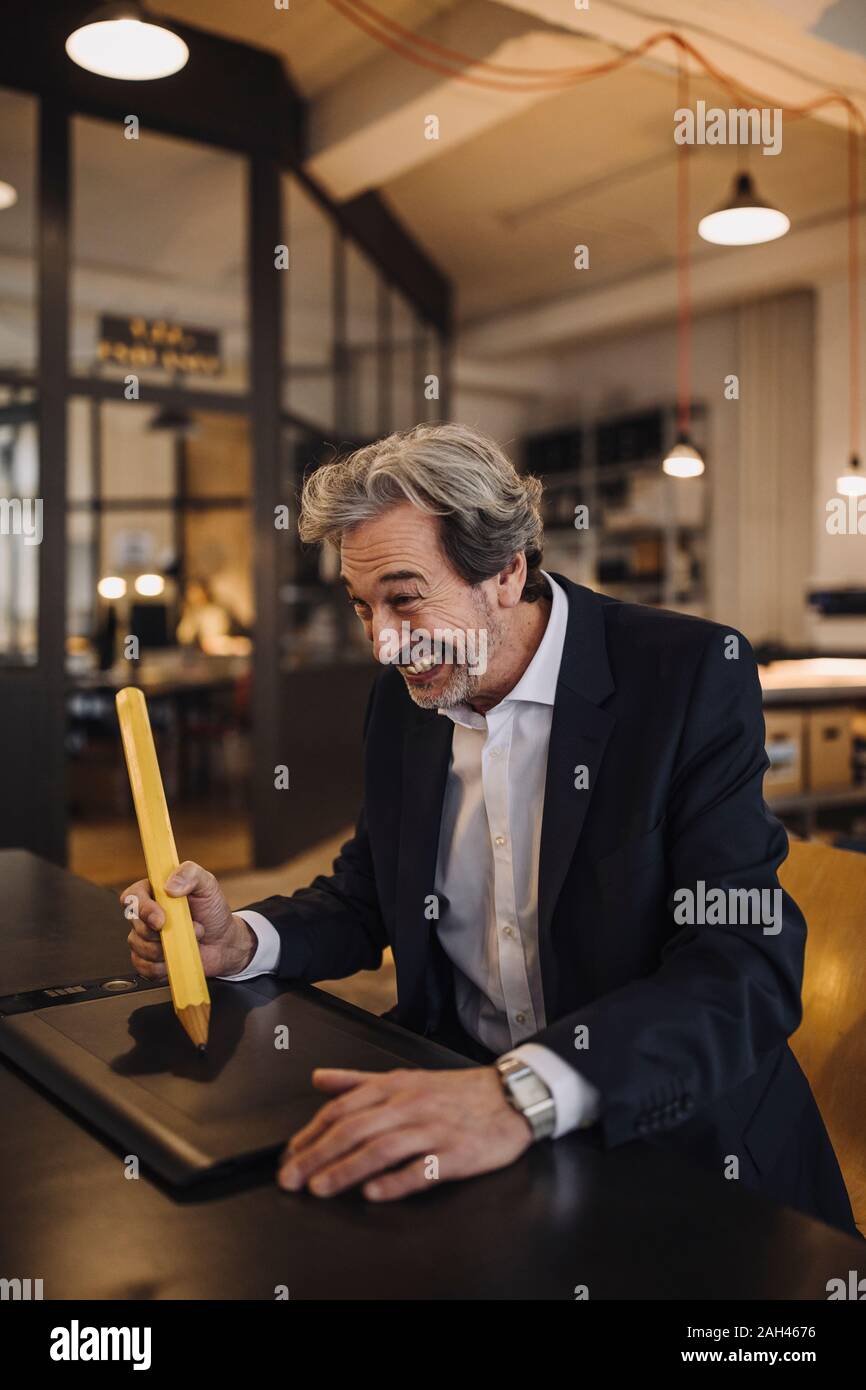 Dumme älterer Geschäftsmann mit riesigen Bleistift am Schreibtisch im Büro Stockfoto