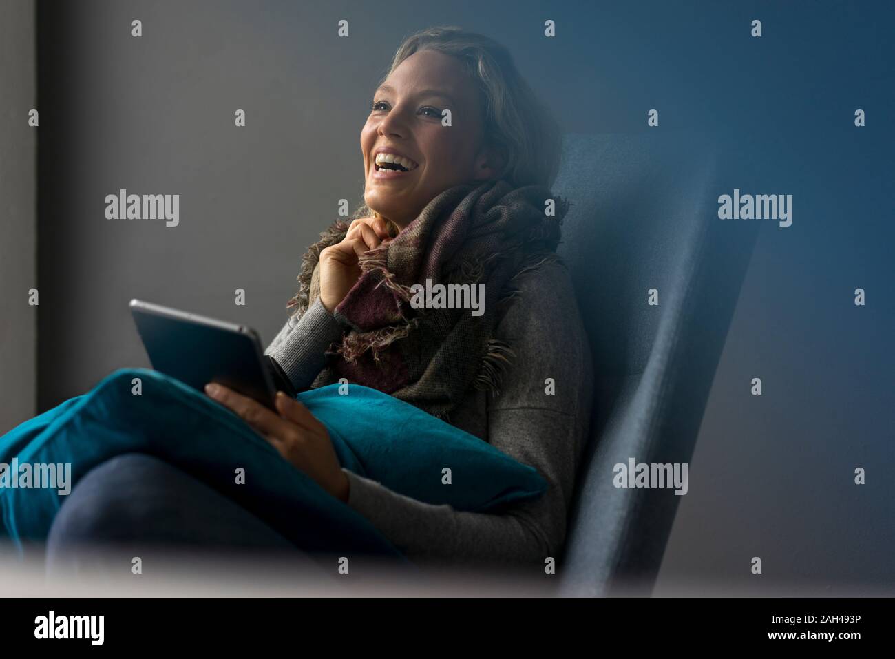 Lachende junge Frau im Sessel sitzt mit Tablet Stockfoto