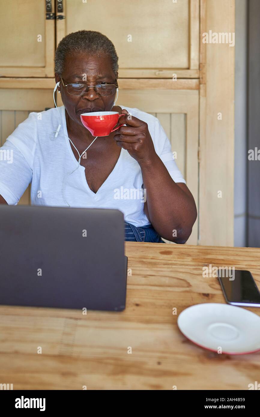 Ältere Frau am Küchentisch sitzen mit Laptop und das Trinken aus dem Becher Stockfoto