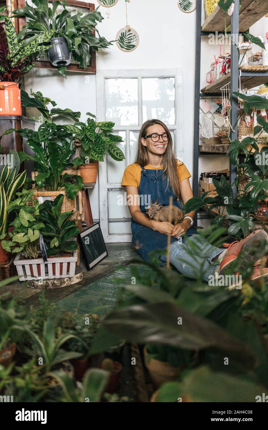Glückliche junge Frau mit Dinosaurier Figur sitzen auf dem Boden in einem kleinen Laden mit Pflanzen Stockfoto