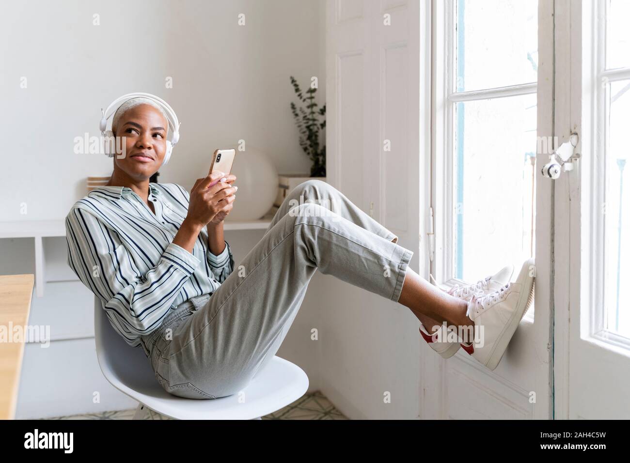 Lächelnde junge Frau mit Kopfhörer und Handy am Fenster Stockfoto