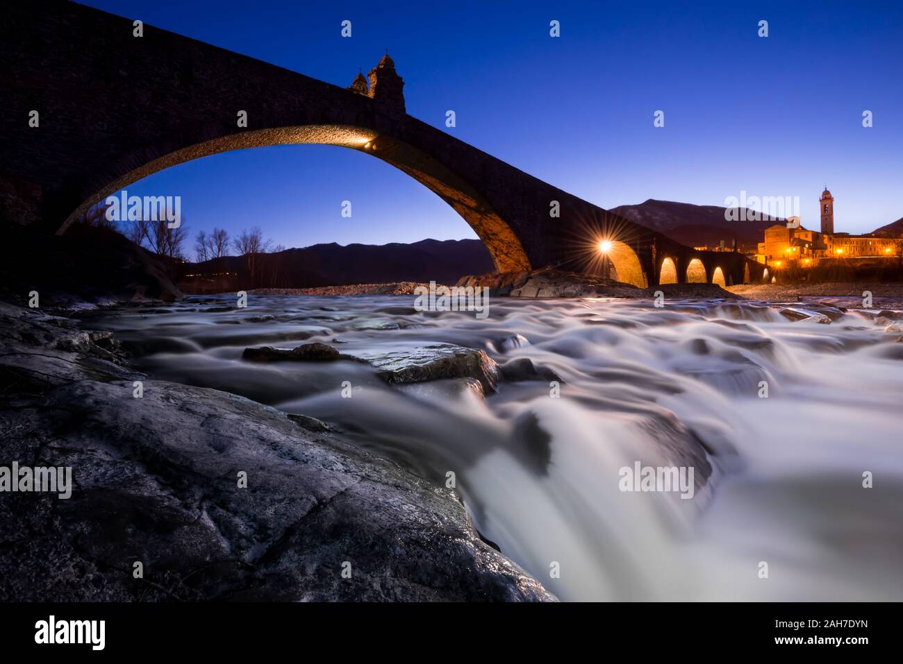 Niedriger Winkel Blick in der Dämmerung einer alten römischen Brücke, die zu einem kleinen italienischen Dorf führt, mit Steinen und einem tobenden Fluss im Vordergrund Stockfoto