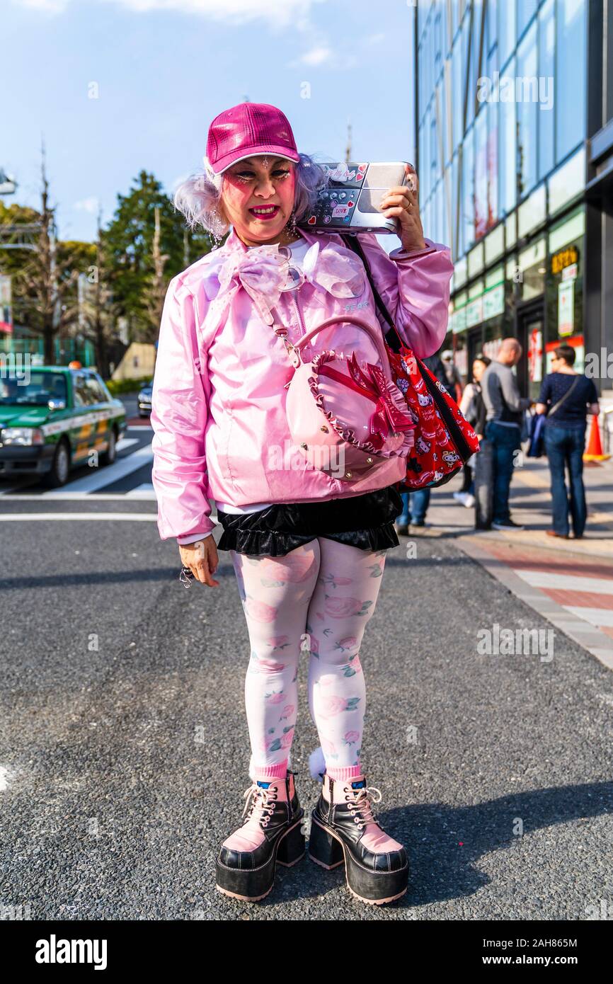 Kawaii Deocra-Chan Japanese Street Fashion. Reife Frau in Rosa mit Hello Kitty Tasche gekleidet. Lächelnd und 2 Finger Frieden Geste geben. Harajuku. Stockfoto