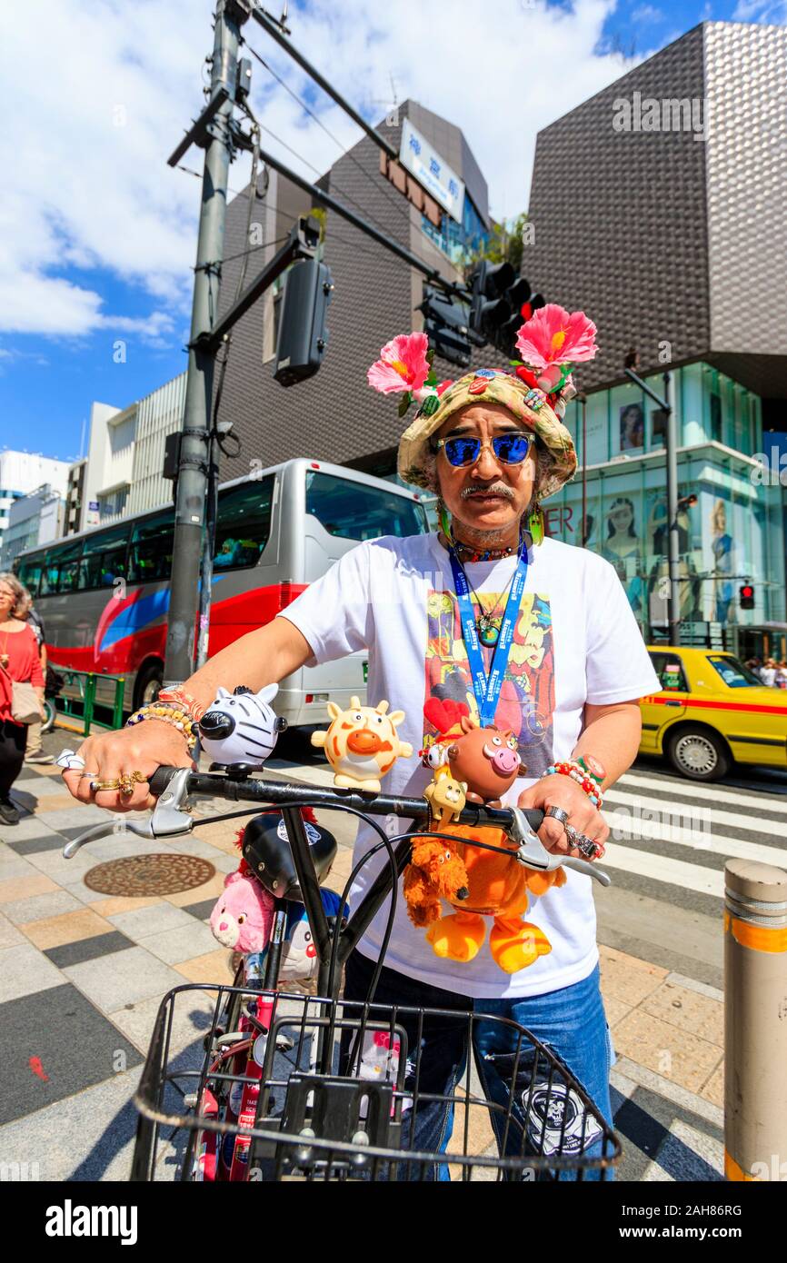 Reifer Mann, Japanisch, Blickkontakt. Stehend mit Fahrrad für Viewer posiert, tragen Sonnenhut mit Blumen und Abzeichen, und viele Freundschaftsbänder. Stockfoto