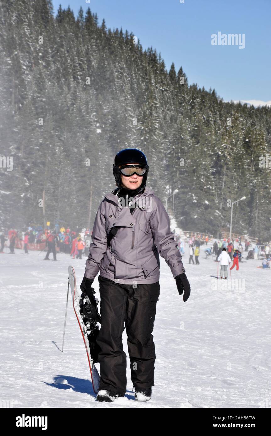 Bansko Bulgarien Ski und Snowboarden lächelnde Snowboarderin hält Snowboard und geht an einem schönen sonnigen Tag auf der Skipiste. Stockfoto