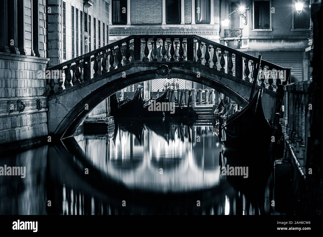 Ikonische schwarz-weiße Nachtaufnahme eines venezianischen Kanals mit Gondeln über Wasser und einer Marmorbrücke, die sich auf dem Wasser spiegelt Stockfoto