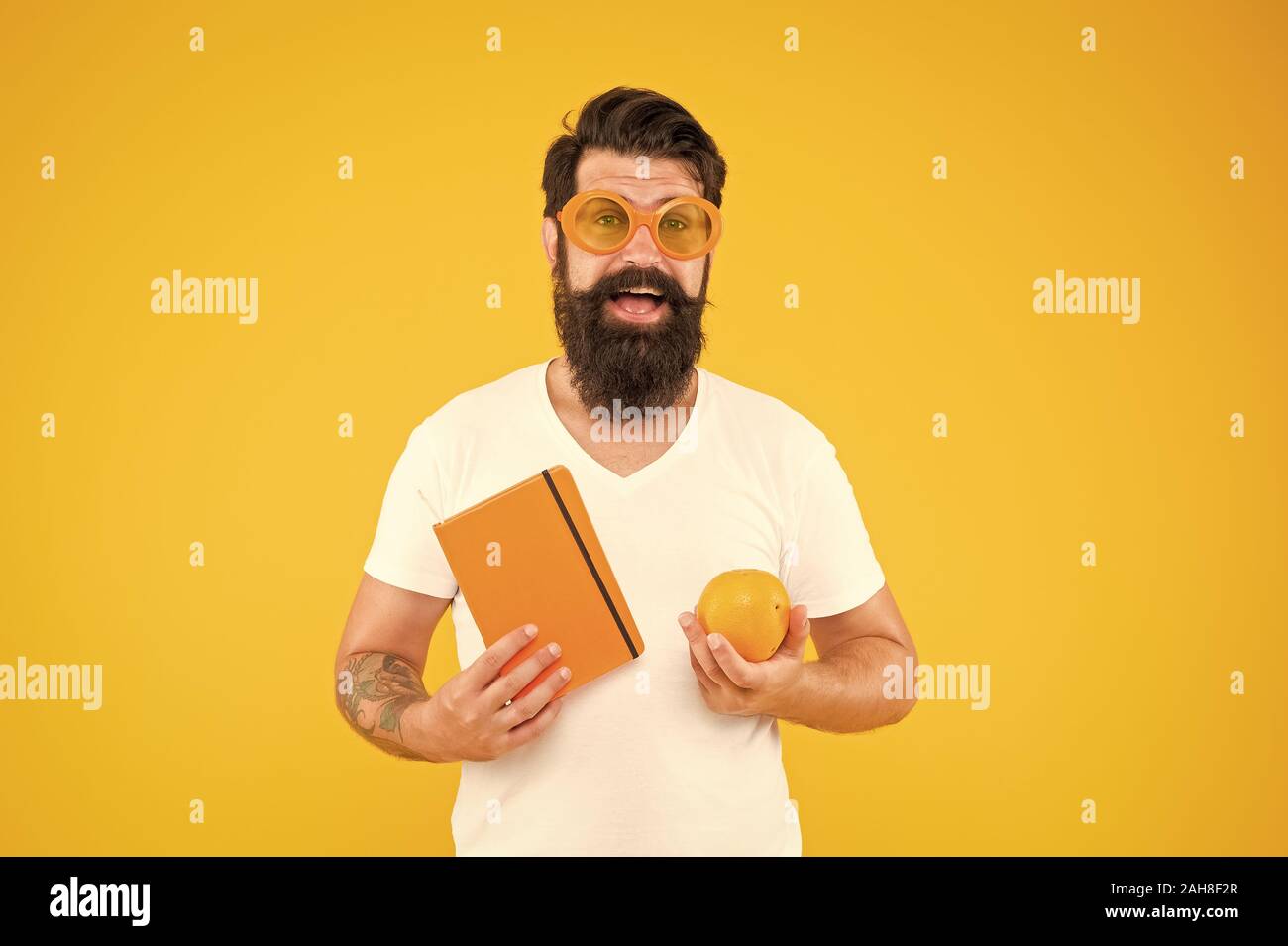 Stolz der geek in Sie. Geek Mann. Bärtiger Mann in geek Brille Holding orange und Buch auf gelben Hintergrund. Hipster in Geek Chic style Auswahl gesunder Nahrung für das Gehirn. Stockfoto