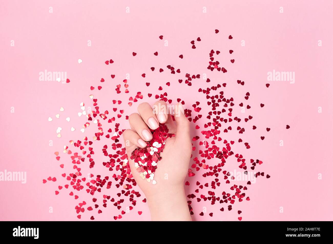 Schöne weibliche Hand, die roten Folie Herz mit leuchtendem Rot funkelnden Herzförmiges Konfetti auf rosa Hintergrund gegossen. Flach. Liebe und Celebratio Stockfoto