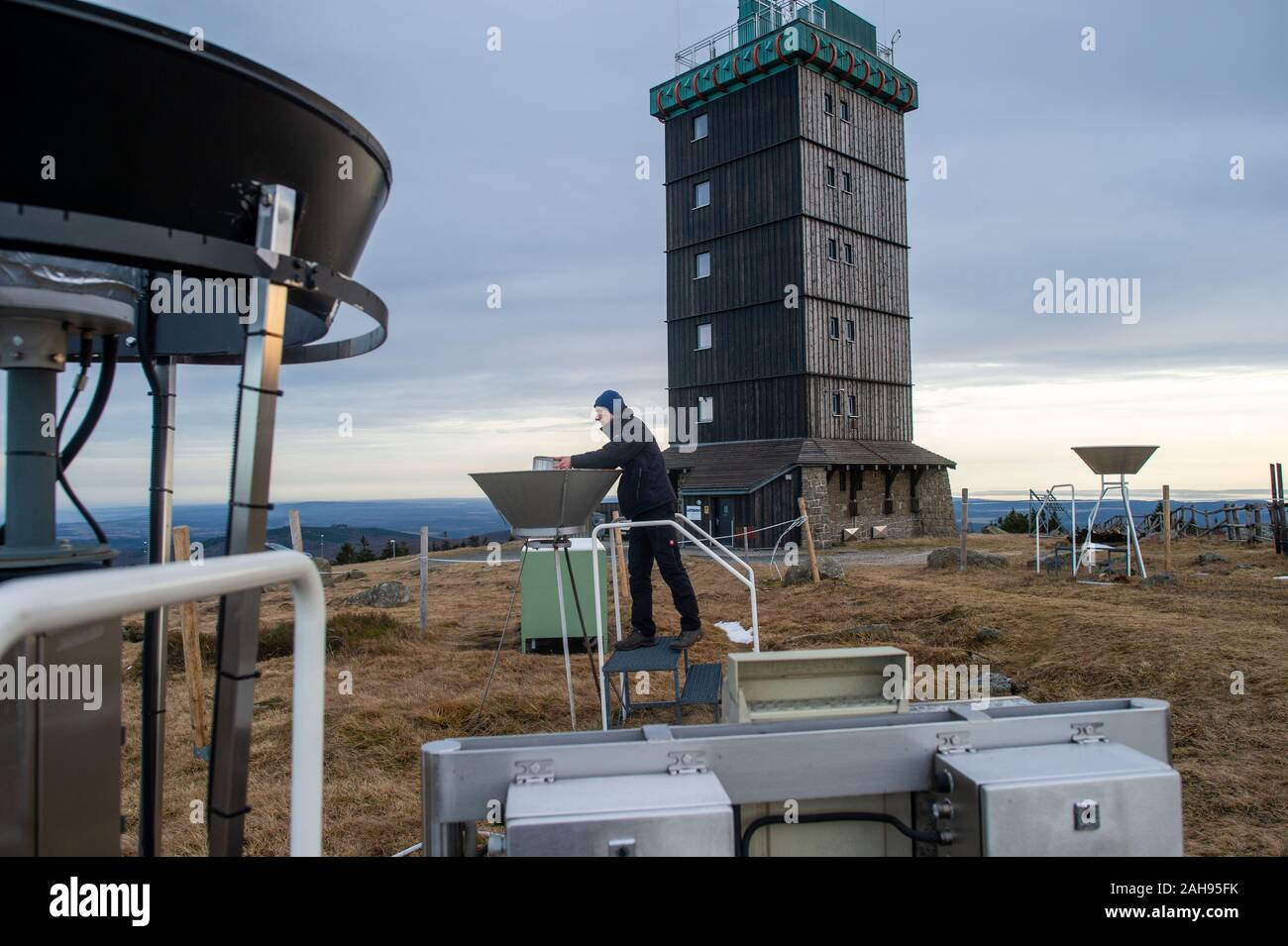 19 Dezember 2019, Sachsen-Anhalt, Wernigerode: Marc Kinkeldey, Wetter beobachter des Deutschen Wetterdienstes (DWD) prüft eine manuelle Messstation auf dem Brocken die Menge des Regens zu bestimmen. Im Vordergrund sehen Sie den automatisierten Messplatz für Regen. Seit 180 Jahren den Brocken war eine extreme Arbeitsplatz für Wetter Beobachter. Ab dem 1. Januar 2020, automatische Messtechnik übernehmen. Wie bei anderen Gipfel wie die Zugspitze und dem Fichtelberg, der Deutsche Wetterdienst ist jetzt stoppen, die Arbeit der Wetter Beobachter auf dem höchsten Gipfel des Harzes. Im nächsten Jahr, nur Radioaktivität Stockfoto