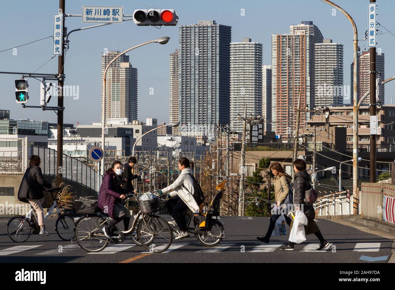 Menschen gehen oder schieben Fahrräder über eine Kreuzung in der Nähe von Shin Kawasaki Station mit den Türmen von Musashi Kosugi hinter.. Kawasaki, Kanagawa, Japan. Stockfoto