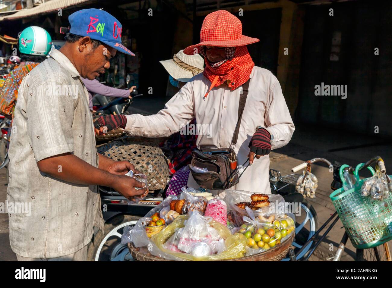Ein asiatischer Mann kauft Lebensmittel von einer Frau verkaufen Obst, Wüste und Donuts zu Ihrem Fahrrad auf einer Straße in Kampong Cham, Kambodscha festgeschnallt. Stockfoto