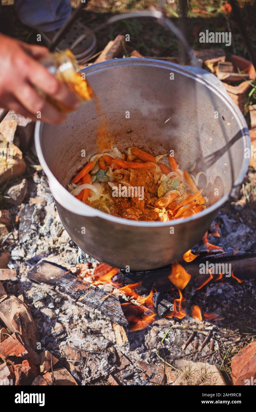 Gießen Gewürze in Gemüse Eintopf. Kochen Abendessen in den großen Kessel auf Camping Feuer. Sonnigen Tag Stockfoto