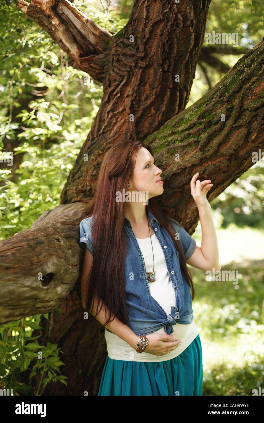 Junge schöne schwangere Frau, die in der Nähe von Tree im Freien Stockfoto