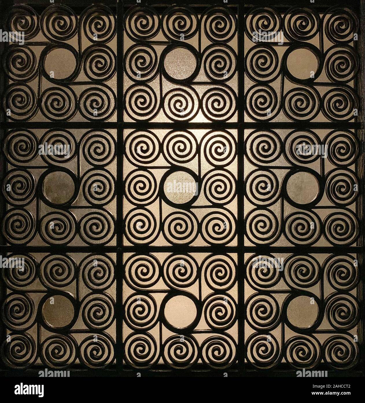 Griechische Wrought Iron Gate Gitter, verziert mit kunstvoll verdrillte Spiralen und mit Milchglas im Hintergrund, dass leicht beleuchtet wird. Stockfoto
