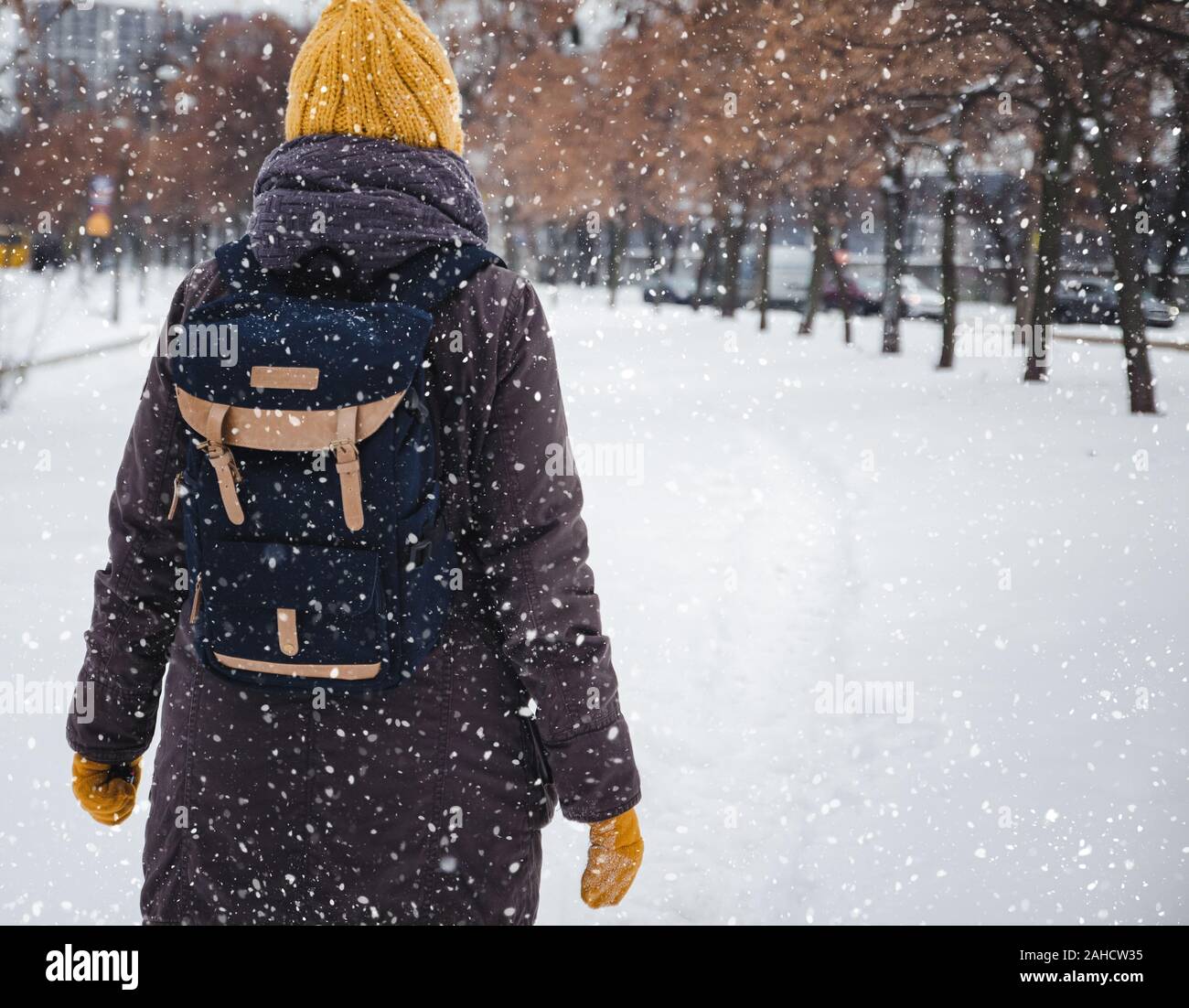 Glückliche junge europäische Frau in einem gelben Strickmütze und Fäustlinge mit einem Rucksack, lacht und lässt unter dem Schneefall. Ansicht von der Rückseite. Retro Effekt Stockfoto