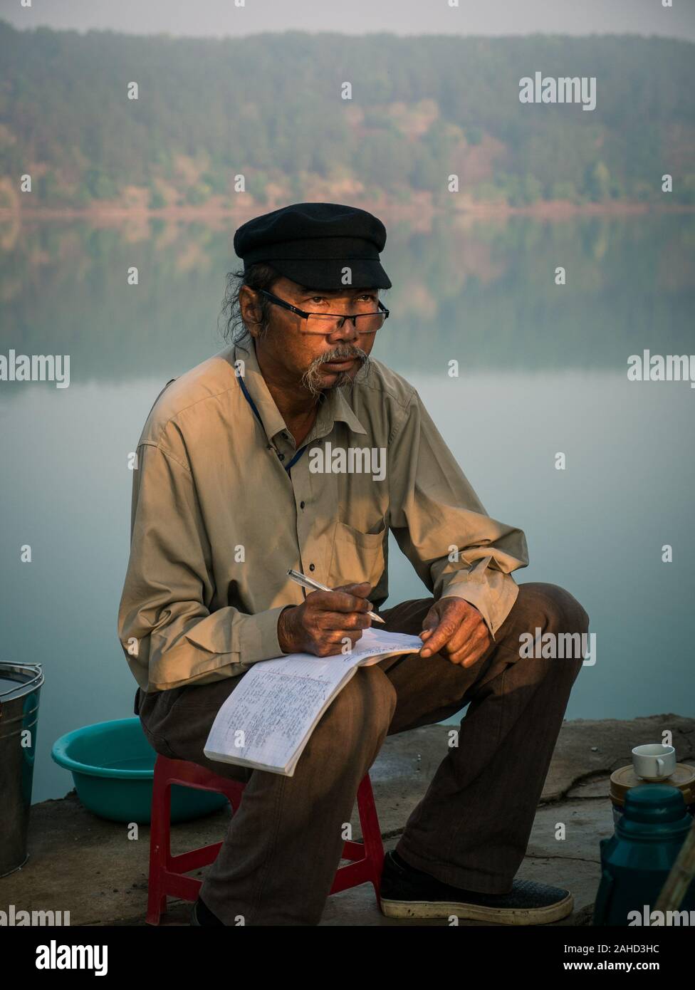 Kreativer vietnamesischer Schriftsteller mit Stift und Buch in der Hand, der mit Sonnenstrahl im Gesicht am See siedet Stockfoto