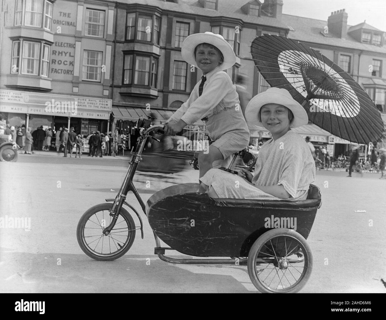 Vintage spätviktorianisches oder frühes Schwarz-Weiß-Foto von Edwardian, das einen jungen Jungen zeigt, der einen großen weißen Hut trägt, mit seiner Schwester ein Fahrrad trittend, einen Sonnenschirm oder einen Regenschirm hält und in einem Beiwagen sitzt, der am Fahrrad befestigt ist. Foto in Rhyl, nordwales. Stockfoto