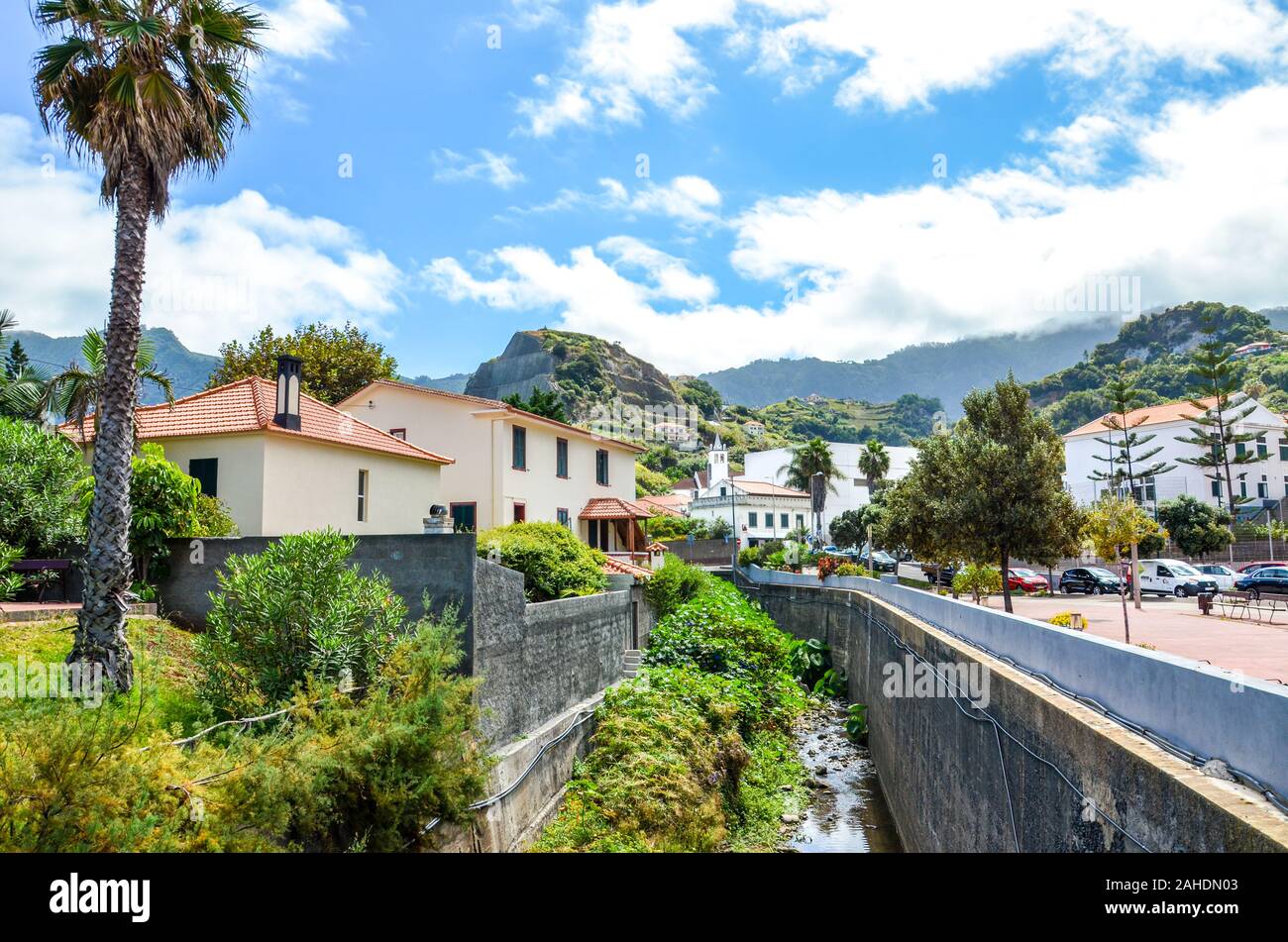 Porto da Cruz, Madeira, Portugal - Sep 24, 2019: Malerische portugiesischen Dorf, das von Hügeln und grünen Vegetation umgeben. Felsen hinter den Häusern. Strom entlang der Straße. Stockfoto