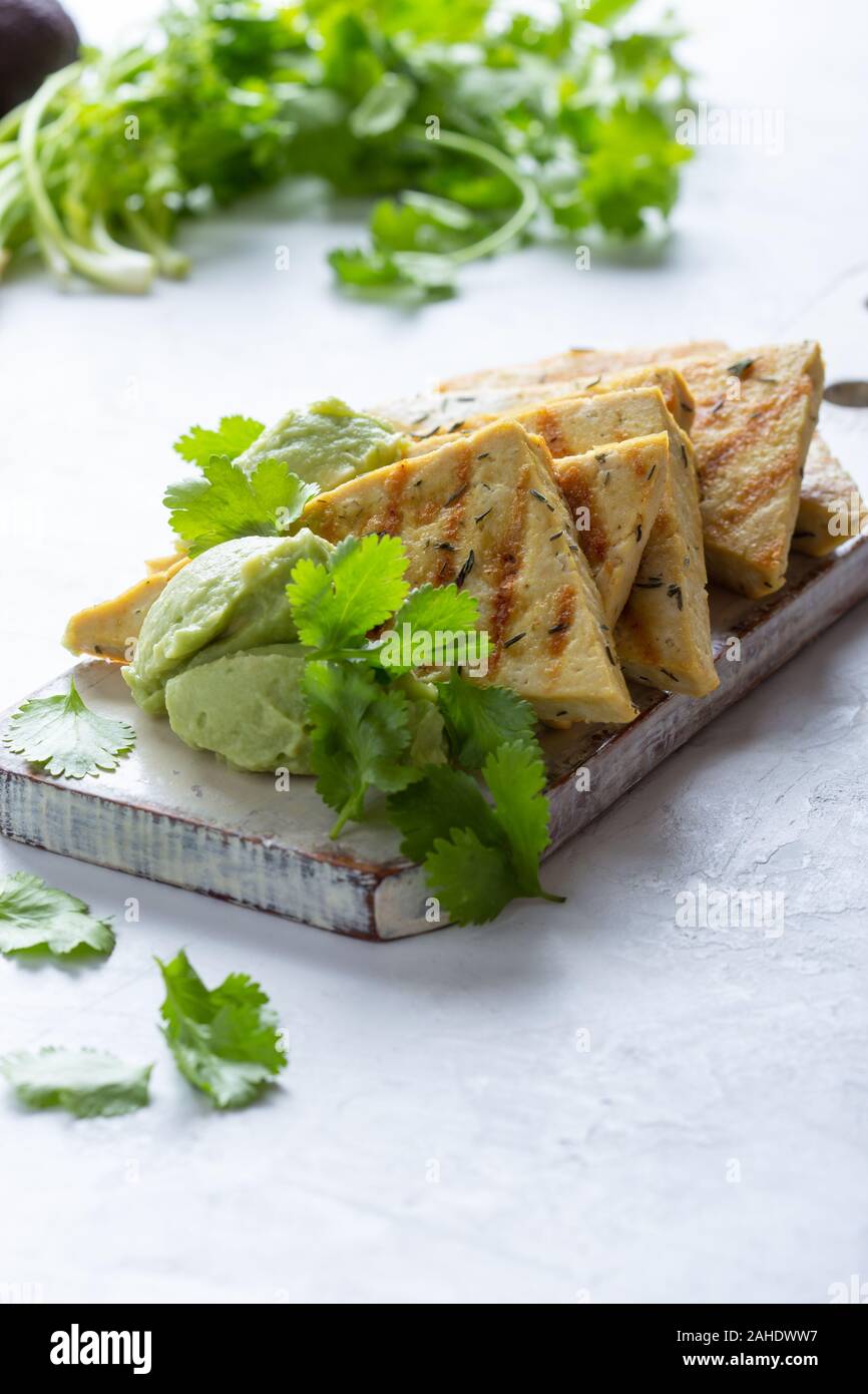 Gegrillte herbed Tofu mit Avocadocreme, vegane Mahlzeit serviert auf rustikalen hölzernen Schneidebrett mit frischem Koriander Kräuter Stockfoto