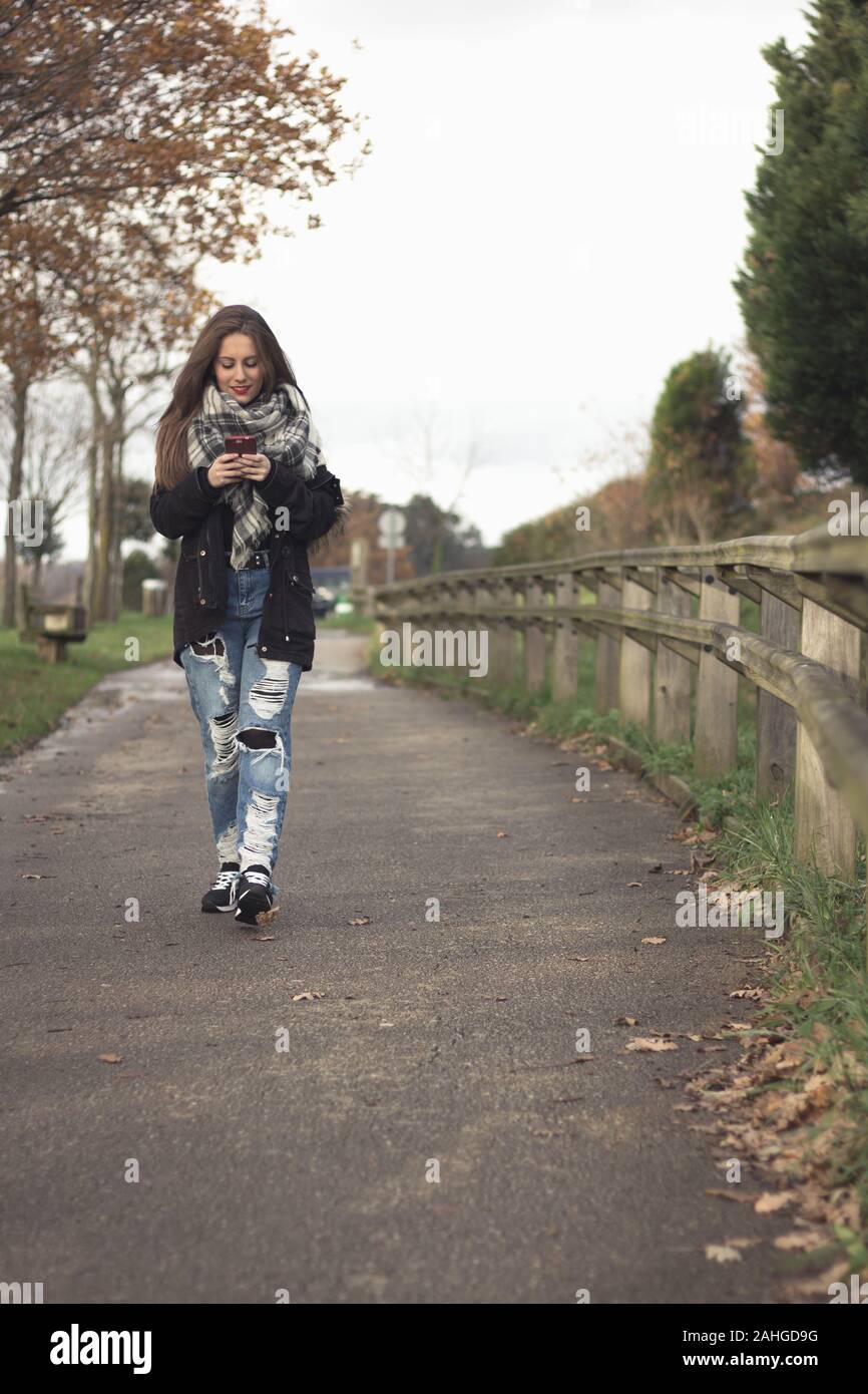 Junge Mädchen tragen zerrissene Jeans Spaziergänge im Park beim Mobiltelefon im Herbst Saison bewölkten Tag suchen. Millenniums-generation cellphone Überbeanspruchung Stockfoto