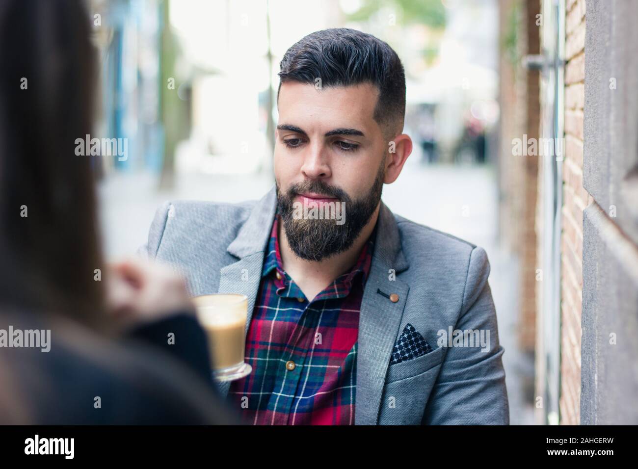 Ein gutaussehender Geschäftsleute, der auf der Café-Terrasse in der Stadt auf ein Kaffee-Glas blickt. Modell mit GanzBarthippchen auf grauem Blazer, der von Cappuccino-Becher begeistert ist Stockfoto