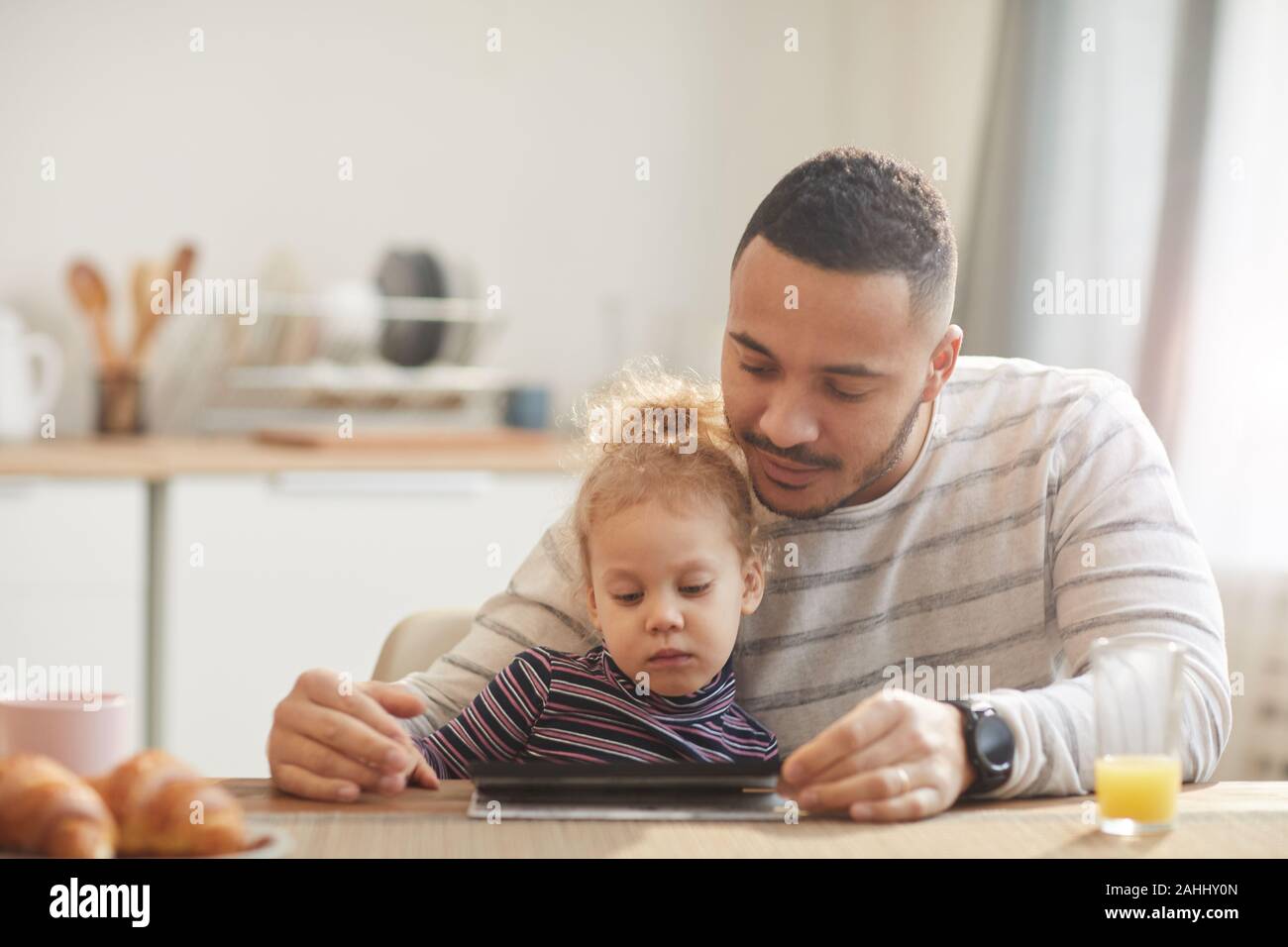 In warmen Tönen Porträt des fürsorglichen Vaters mit niedlichen kleinen Mädchen mit digitalen Tablet zusammen beim Sitzen an den hölzernen Tisch in der gemütlichen Küche, Kopie Raum Stockfoto