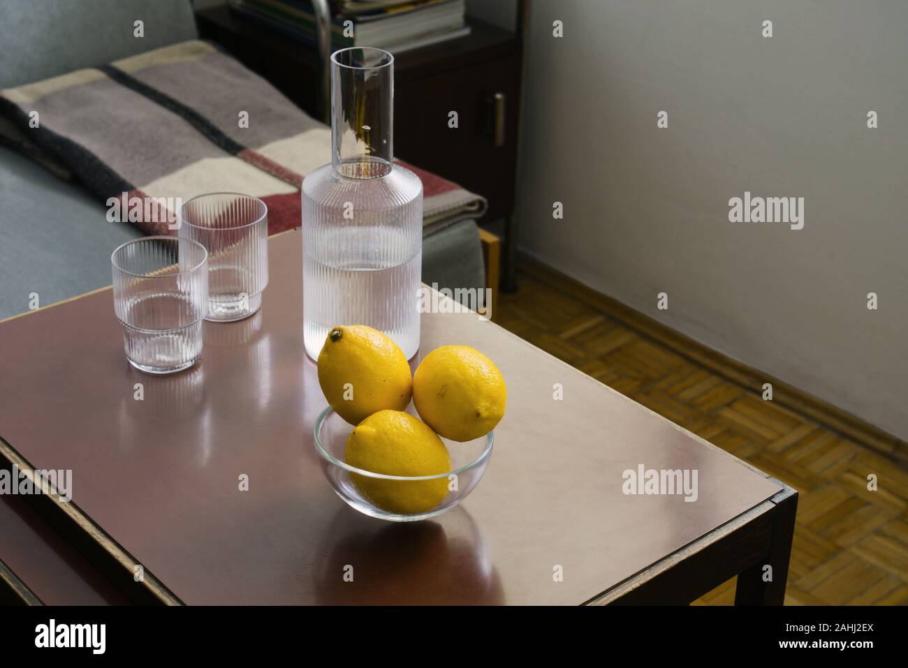 Drei gelbe Zitronen in ein Glas Schale, zwei Gläser und Karaffen mit Wasser auf den Tisch, im Vordergrund konzentrieren, Zimmer Ecke mit Möbel unscharf im Hintergrund Stockfoto