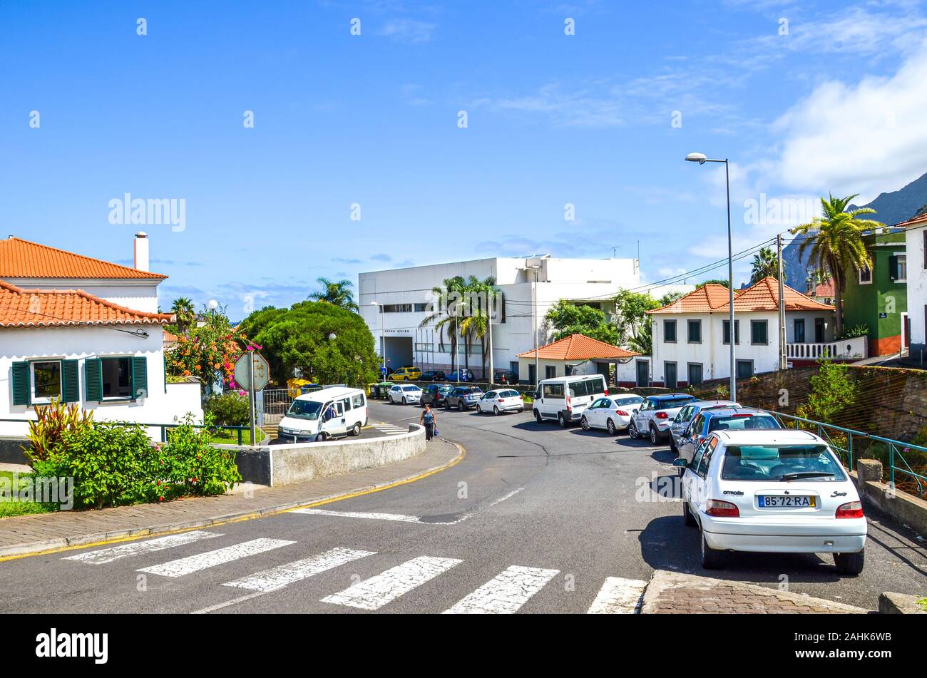 Porto da Cruz, Madeira, Portugal - Sep 24, 2019: Straße der malerischen portugiesischen Dorf. Häuser, Menschen auf der Straße, geparkte Autos. Sonnigen Tag, blauer Himmel. Zebrastreifen im Vordergrund. Stockfoto