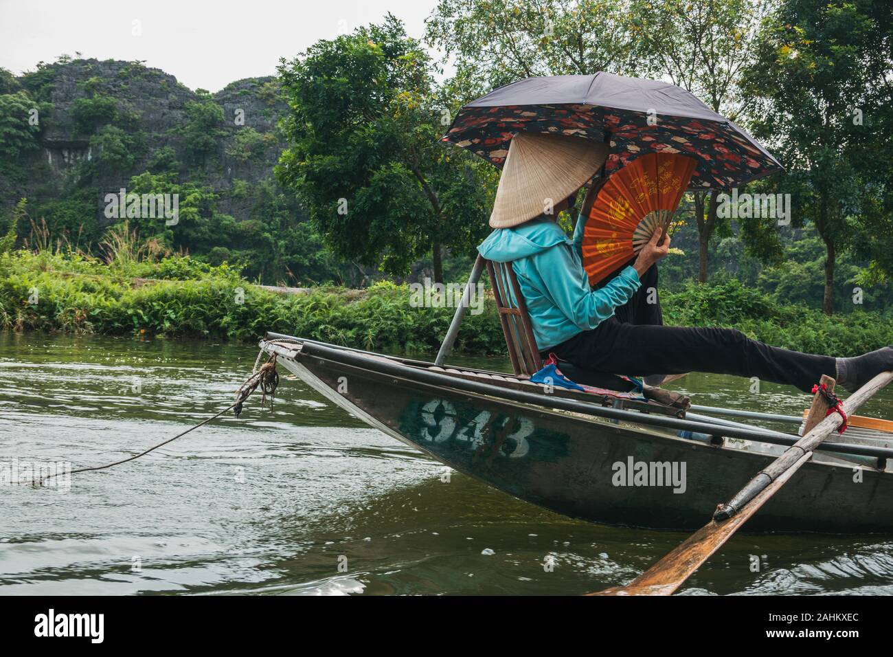 Eine ältere Frau mit ihren Füßen die Ruder der ein Ruderboot zu drücken, während Sie einen Ventilator und Regenschirm, die Touristen den Fluss hinunter in Hoa Lư, Vietnam Stockfoto