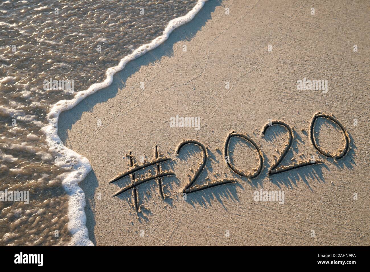 Modernes neues Jahr Nachricht mit social media Hashtag für 2020 handschriftlich in sanften Sand mit Sunrise lens flare über entgegenkommende Welle auf den Strand Stockfoto