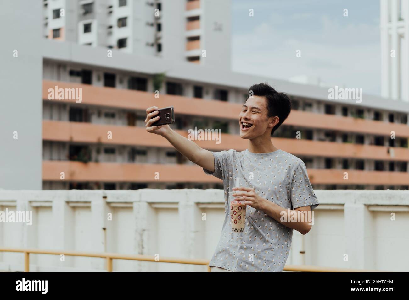 Asiatischer männlicher Vlogger mit Bubble Tea und Smartphone, HDB-Gehäuse im Hintergrund, Content Creator Stockfoto