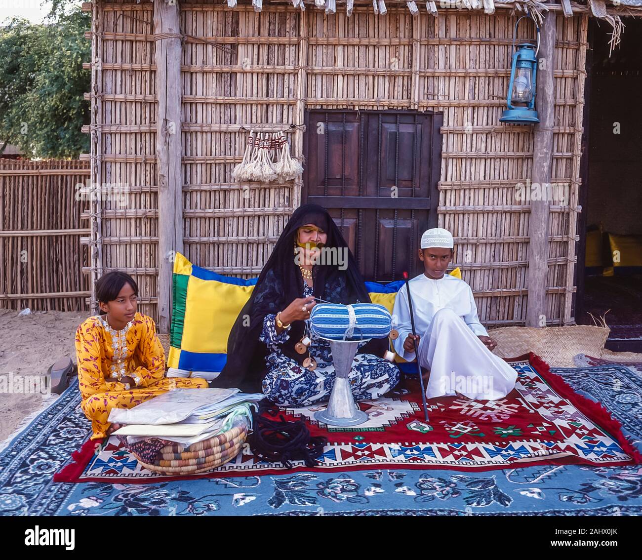 Dubai arabische Folklore und Geschichte mit einem Beduinen emiratische Dame mit ihrer Tochter und Sohn sitzen, während Sie die Fähigkeiten des Webens mit silbernem Faden für den Einsatz auf ceromonial Kleider auf der Shindagha Heritage Village an der Mündung des Dubai Creek während des Dubai Messe und Festival in den Vereinigten Arabischen Emiraten demonstriert entfernt Stockfoto