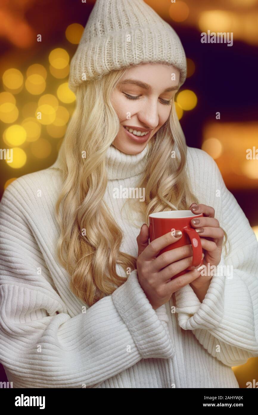 Junge Frau mit einer Tasse heißen Kaffee im Winter schröpfen in ihren Händen, wie sie die Getränke mit einem glückseligen Lächeln gegen einen leuchtenden warmen Orange backgroun Stockfoto