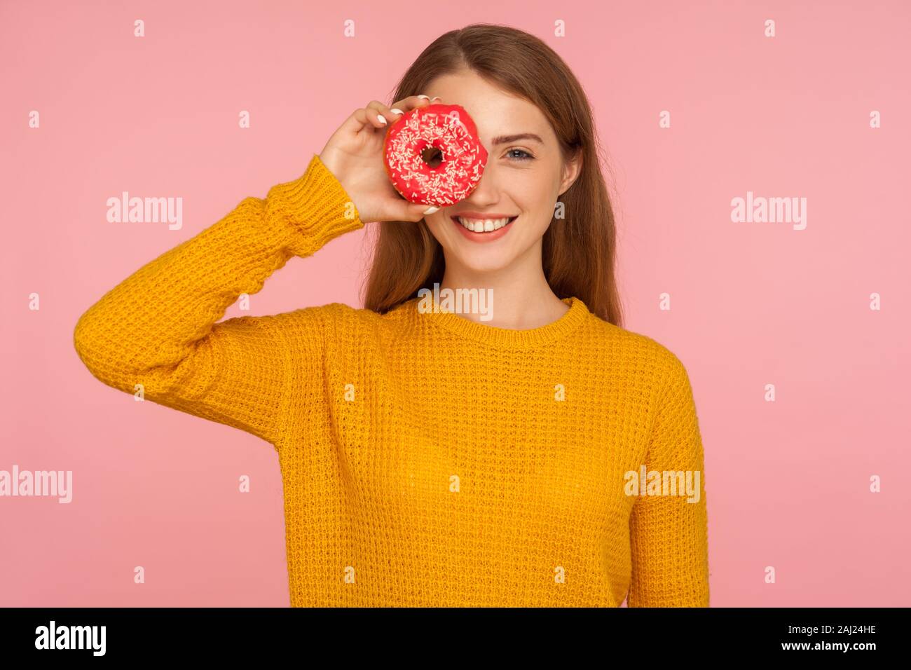 Porträt der schöne Rote Haare Mädchen in Pullover mit Kamera durch die Bohrung in der Donut und lächelnd, spähen in rosa Donut, Spaß haben, spielen wi Stockfoto