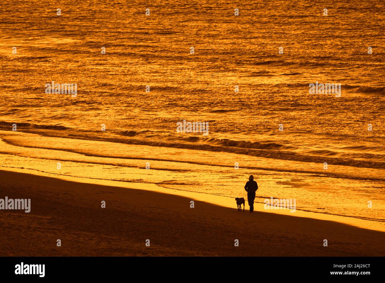 Einsame Hundebesitzer zu Fuß entlang der Küste mit unleashed Hund am Strand, bei Sonnenuntergang Silhouette an einem kalten Abend im Winter Stockfoto