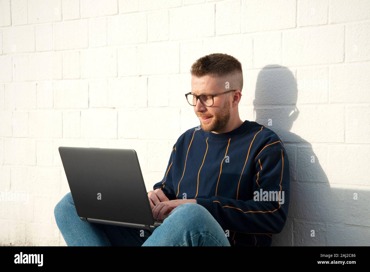 Jungen kaukasischen Mann in casual-wear mit überkreuzten Beinen auf dem Boden und Arbeiten am Laptop. Freelancer arbeiten außerhalb der Texteingabe auf Laptop Computer. Weiß w Stockfoto