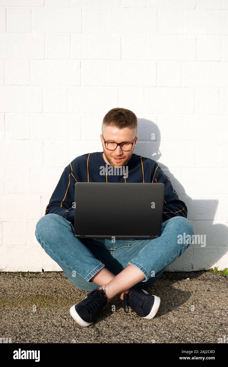 Lächelnd kaukasischen digitale Arbeitnehmer im casual-wear mit überkreuzten Beinen auf dem Boden und Arbeiten am Laptop. Junge Freiberufler arbeiten außerhalb der Texteingabe auf lapto Stockfoto