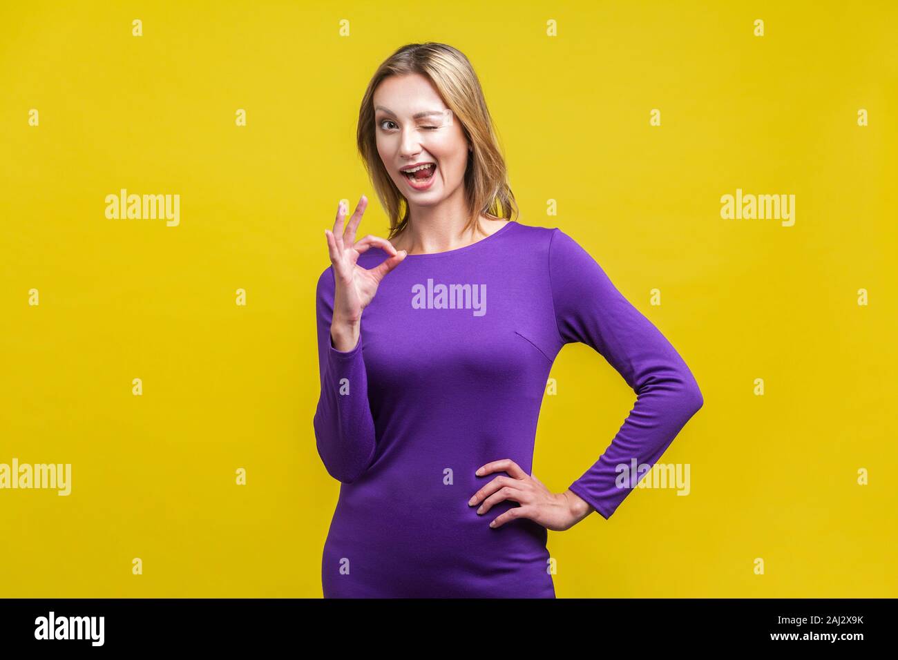 Alles ist in Ordnung. Portrait von verspielten positive Frau im lila Kleid stehend mit einer Hand auf den Hüften, an Kamera winking und zeigen ok Geste. Stockfoto