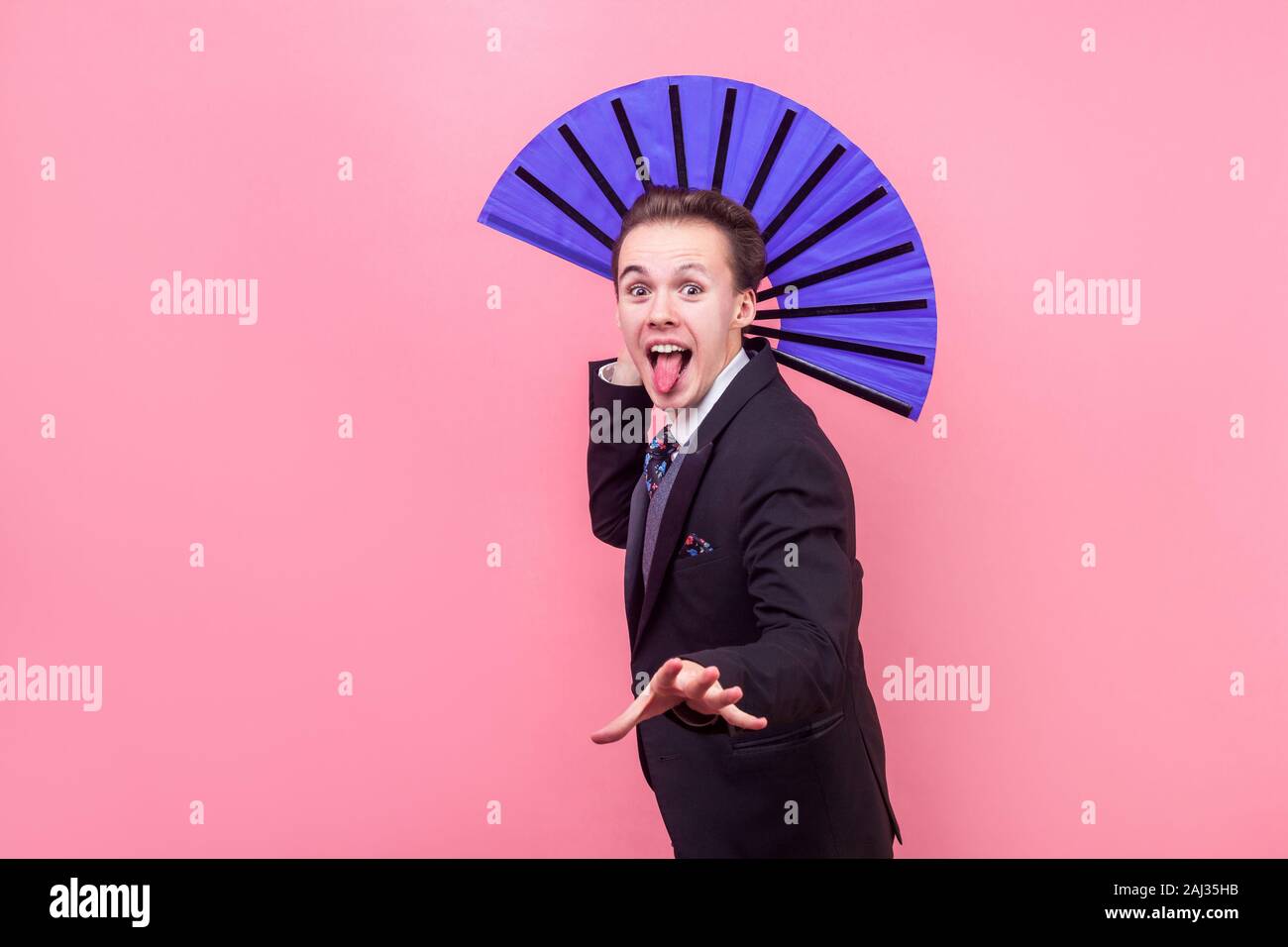 Portrait von fröhlichen unbeschwerten jungen Mann, Zauberer im eleganten Anzug Zunge heraus und halten big blue Ventilator, Illusionist, Leistung, Stockfoto