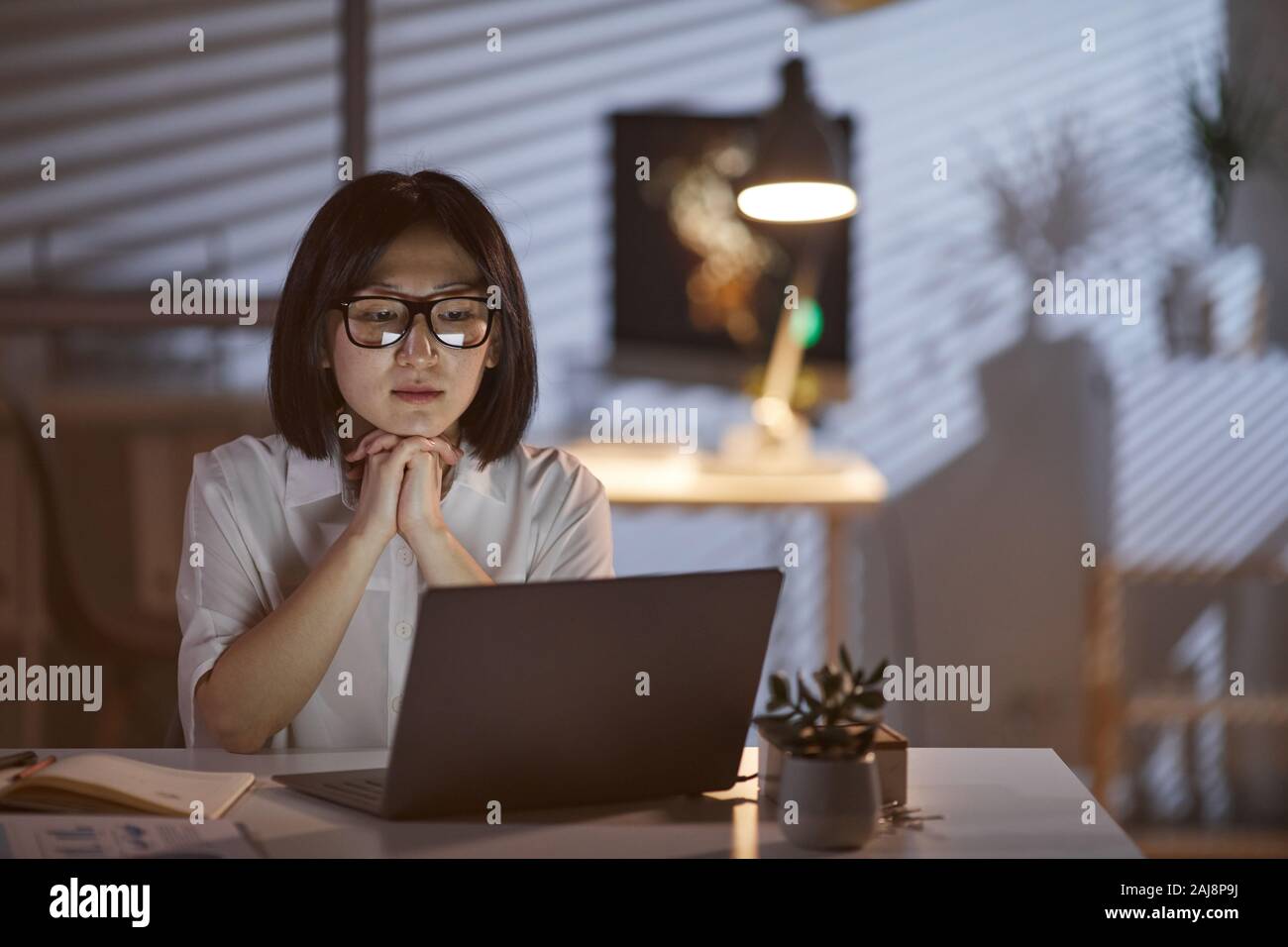 Asiatische junge Frau mit dunklen kurzen Haaren auf Monitor des Laptops mit nachdenklichen Blick, während bei Dunkelheit die Arbeit im Büro Stockfoto