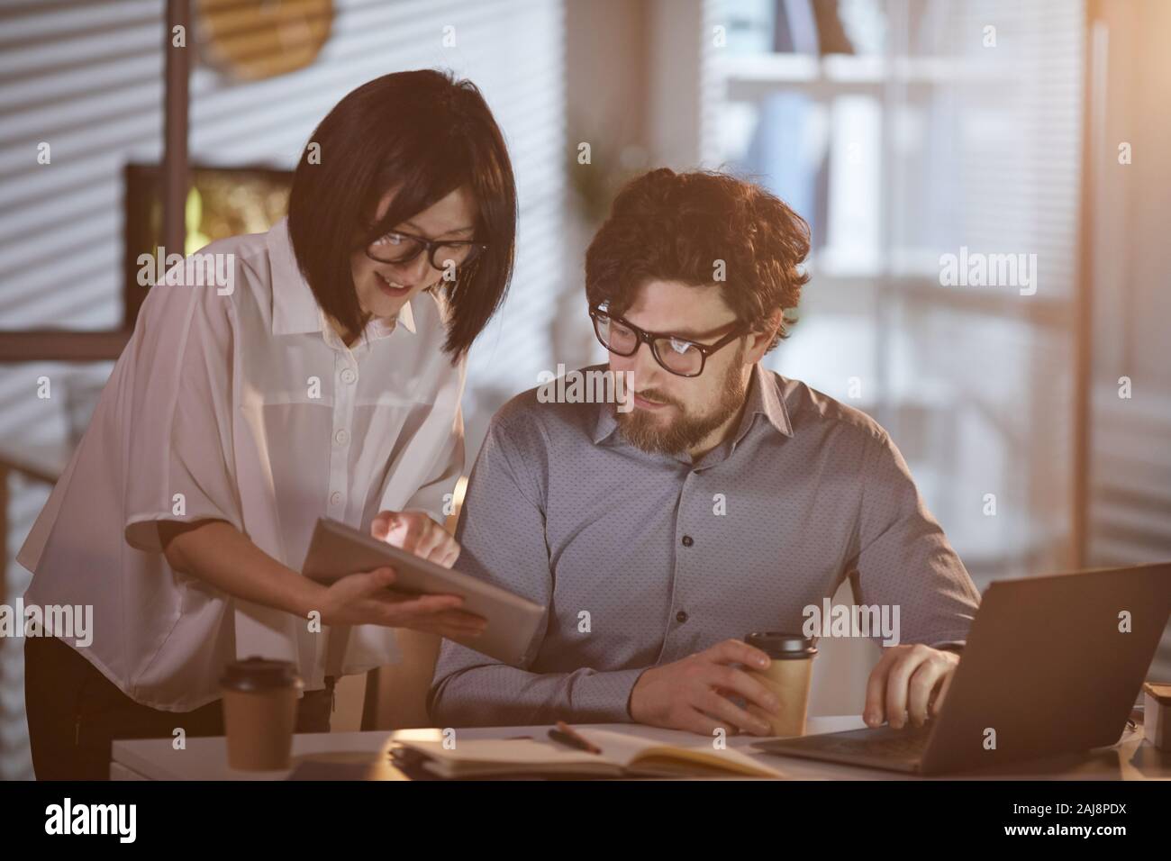 Asiatische junge Geschäftsfrau, die auf digitalen Tablet und etwas mit ihrem Kollegen, während er auf Laptop im Büro arbeiten Stockfoto