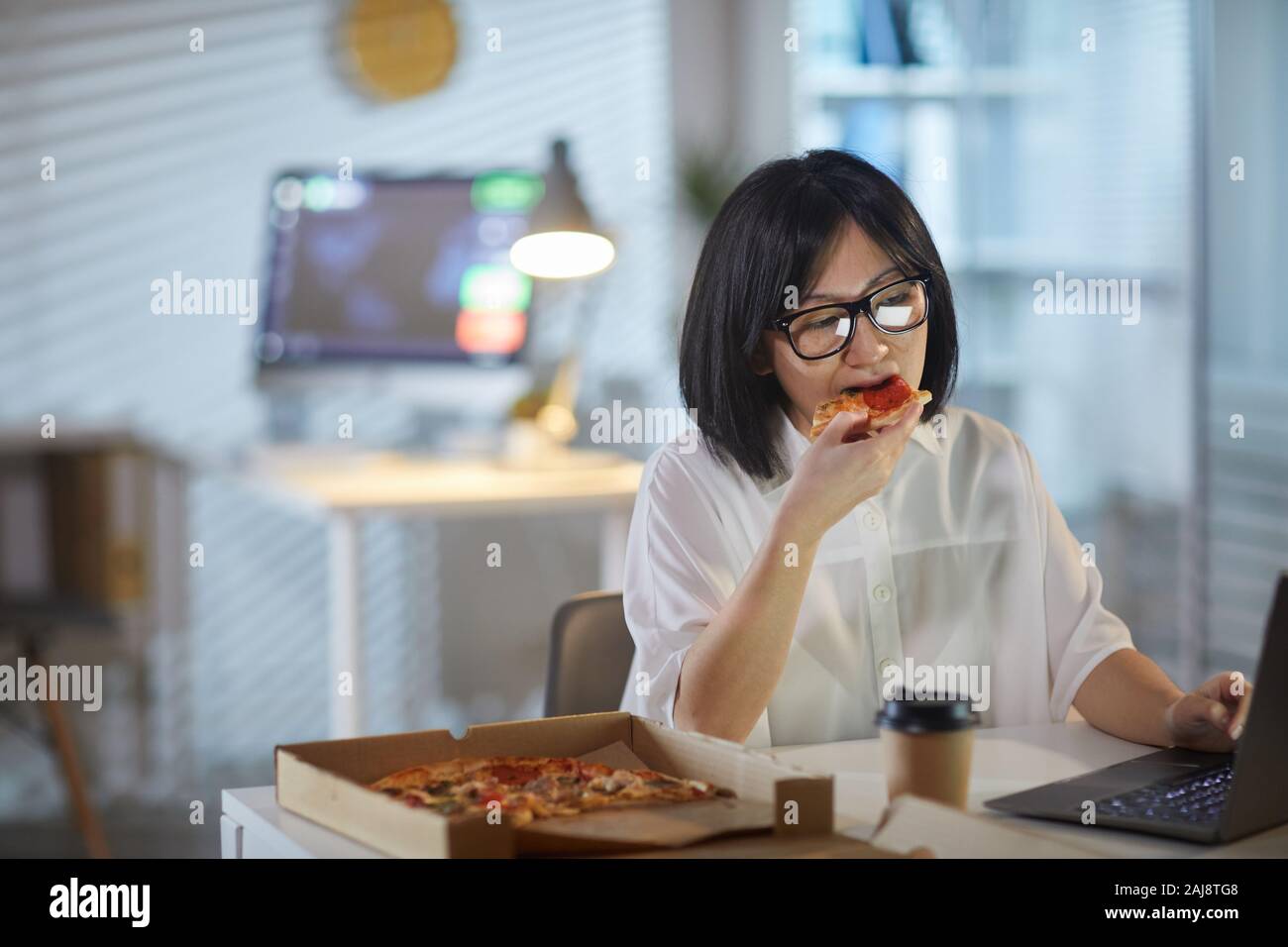 Asiatische Geschäftsfrau beißen ein Stück Pizza und Arbeiten am Laptop am Tisch im Büro Stockfoto