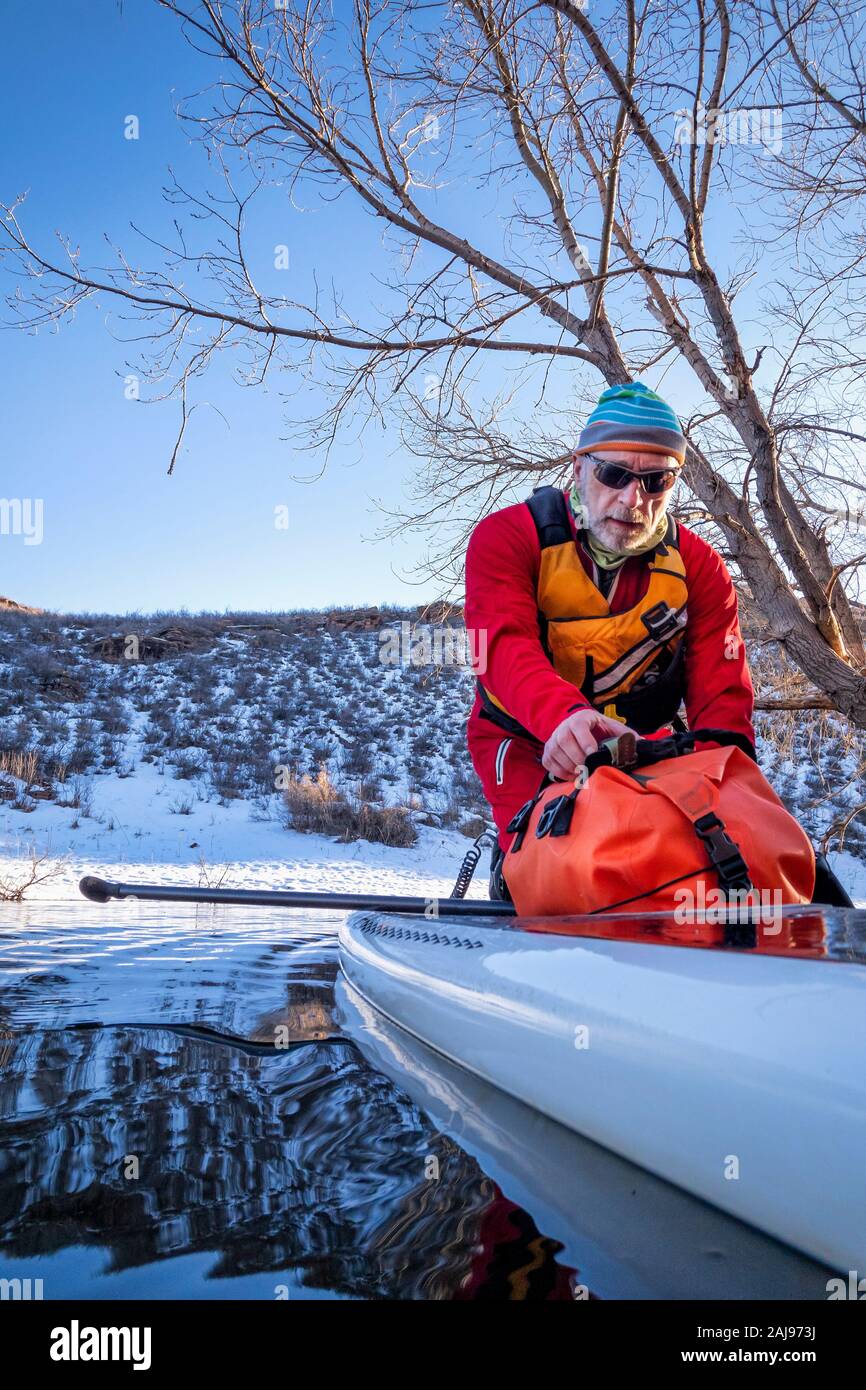 Senior Paddler ist gerade eine trockene Tasche auf einem Deck von Stand up paddleboard auf einem Bergsee im Winter Landschaft - horsetooth Reservoir in Northern Colorado Stockfoto