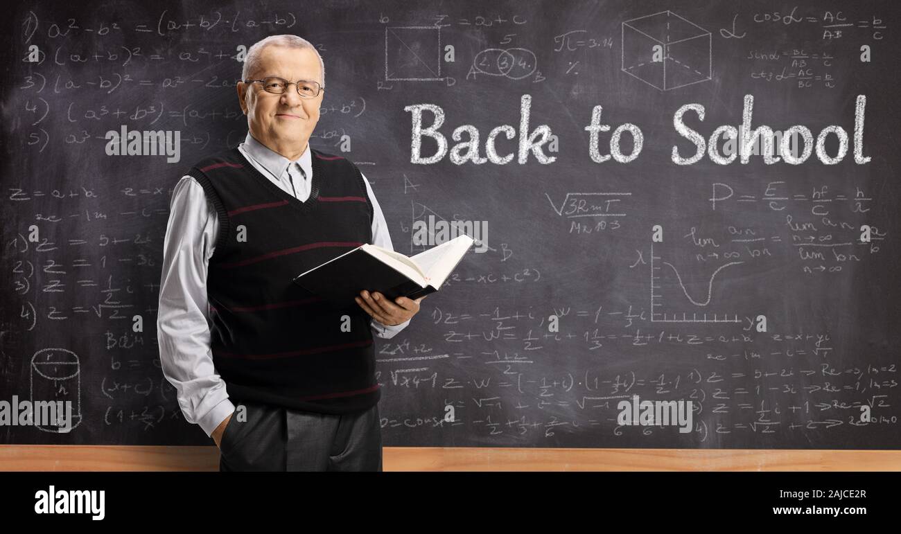 Ältere männliche Lehrer mit, die ein Buch vor einer Schule Tafel geschrieben mit zurück in die Schule text und Mathe forumlas Stockfoto