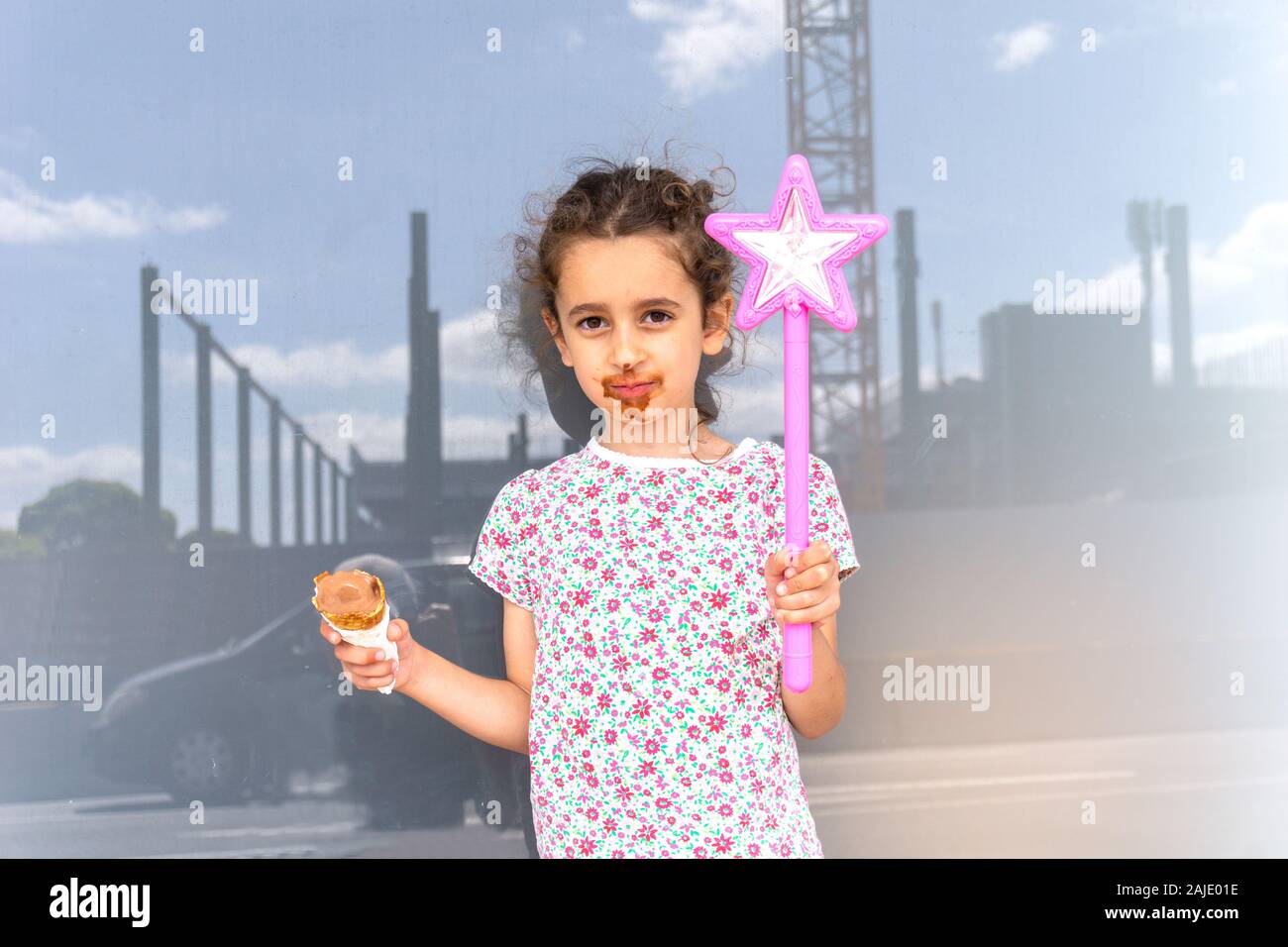 Kleines Mädchen mit unordentlichen Gesicht genießen Sie Schokolade essen halten rosa Zauberstab mit Stern in der Stadt mit dem Bau Skyline spiegelt sich in Fenster hinter. Stockfoto